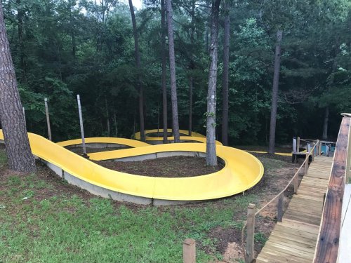 A winding yellow slide surrounded by trees, leading down from a wooden platform in a forested area.