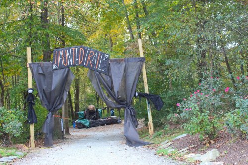 A spooky entrance to a haunted trail, marked by a sign and draped black fabric, surrounded by trees and flowers.
