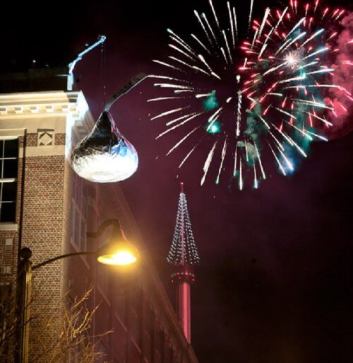 A giant silver kiss-shaped object hangs from a building, with colorful fireworks lighting up the night sky.