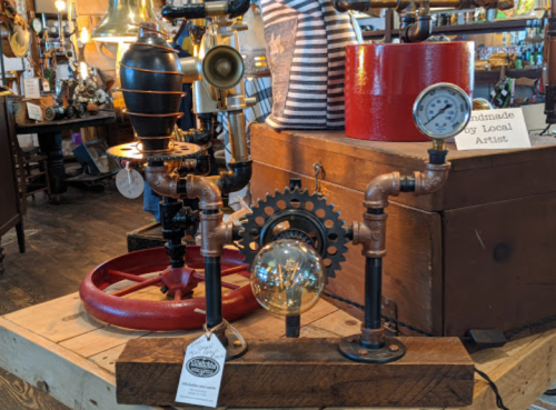 A collection of steampunk-style lamps and decor featuring pipes, gears, and a vintage gauge on a wooden display.