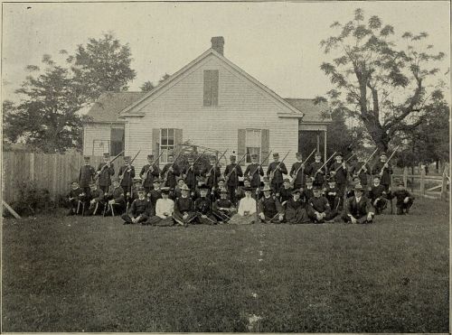 A historical group photo of soldiers in uniform, posed in front of a house with rifles, surrounded by trees.