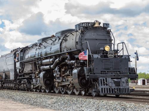 A vintage steam locomotive with an American flag, parked on tracks under a cloudy sky.