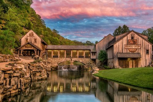 A rustic building by a serene pond, with a waterfall and colorful sunset sky in the background.