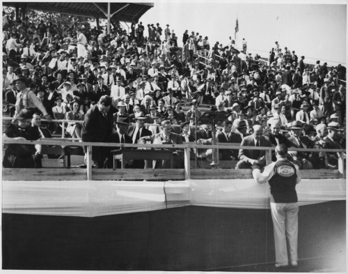 A large crowd seated in a grandstand, with a speaker addressing them from a stage below.
