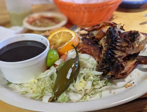 A plate of grilled chicken with black beans, lettuce, jalapeño, lime, and an orange slice, served in a casual dining setting.