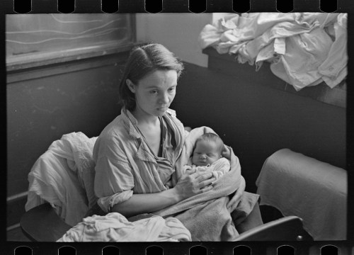 A woman holding a newborn baby, sitting in a room with laundry piled nearby. The atmosphere is calm and intimate.