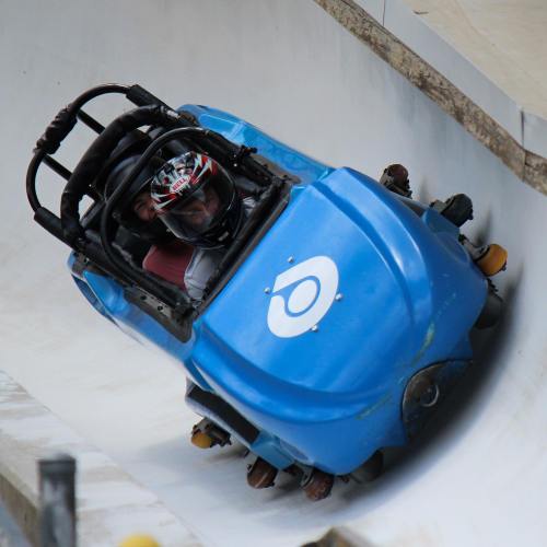 Two people in a blue bobsled racing down a curved track, wearing helmets and focused on the ride.