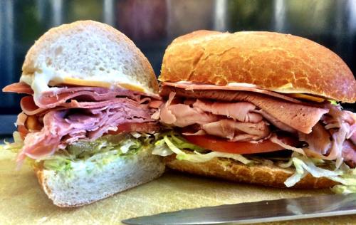 A freshly cut sandwich with layers of ham, turkey, lettuce, tomato, and mayonnaise on a soft roll, placed on a cutting board.