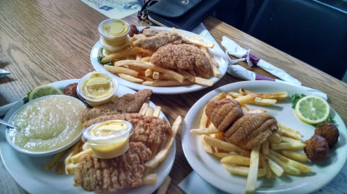 Three plates of fried fish and chicken, served with fries, lemon slices, and dipping sauces on a wooden table.