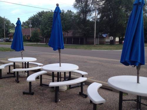 Four outdoor tables with blue umbrellas are set up along a quiet street, surrounded by trees and buildings.