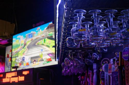 A bar scene with hanging glasses and a colorful video game displayed on a screen in the background.