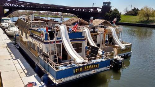 Two colorful houseboats with slides docked by a river, featuring tropical decor and a bridge in the background.