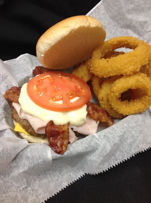 A burger with bacon, tomato, and mayo, served with a side of crispy onion rings in a takeout container.