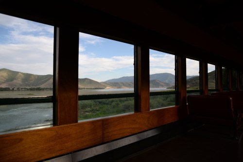 View from a train window showcasing a scenic landscape of mountains and a river under a blue sky.