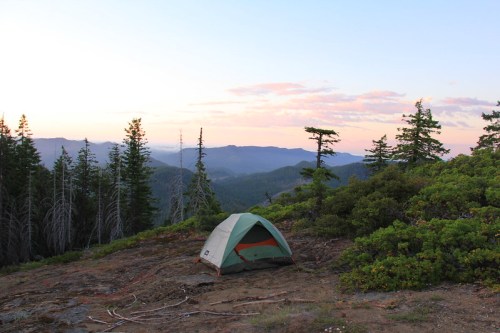 A green tent set on rocky ground, surrounded by trees, with a scenic mountain view at sunset.