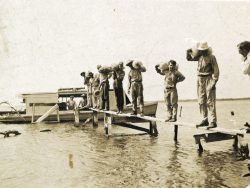 A group of people stands on a wooden dock by a boat, looking out over the water in a vintage black-and-white photo.