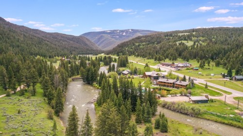 Aerial view of a lush valley with a winding river, surrounded by mountains and dotted with cabins and trees.