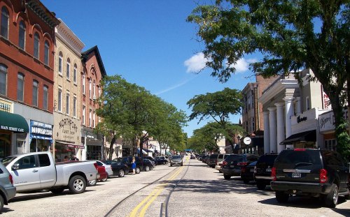 A charming downtown street lined with shops, trees, and parked cars under a clear blue sky.