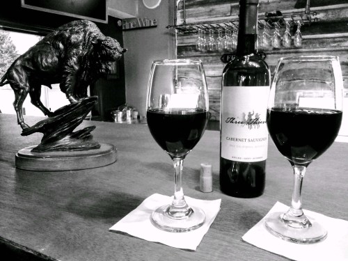 A bottle of Cabernet Sauvignon with two glasses of wine on a wooden bar, alongside a bison sculpture.