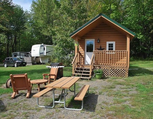 A cozy wooden cabin with a porch, picnic table, and nearby RVs, surrounded by lush green trees.