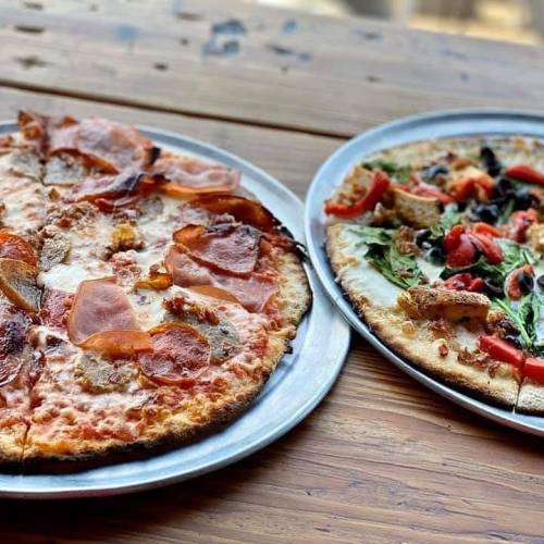 Two pizzas on metal trays: one with meats and cheese, the other topped with vegetables and greens, on a wooden table.