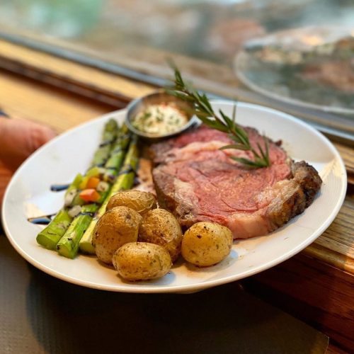 A plate of roasted potatoes, asparagus, and a slice of prime rib with a dipping sauce, served on a wooden table.