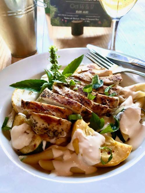 A bowl of pasta topped with grilled chicken, fresh herbs, and a creamy sauce, with a drink and menu in the background.