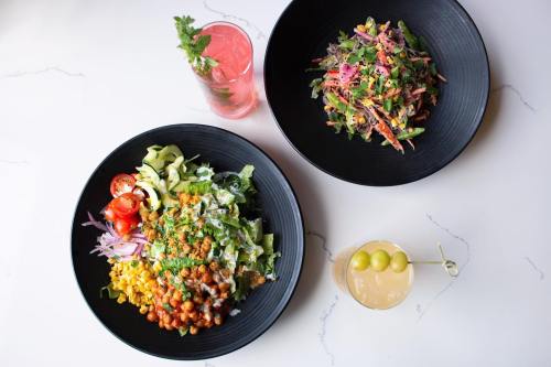 Two black bowls of colorful salads with fresh vegetables, accompanied by vibrant drinks on a marble table.