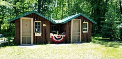 Two rustic wooden cabins with green roofs, surrounded by trees and a grassy area, featuring flower boxes and patriotic decor.