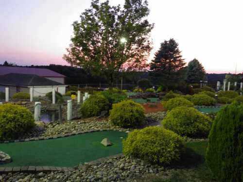 A mini-golf course at sunset, featuring lush greenery, shrubs, and trees under a colorful sky.
