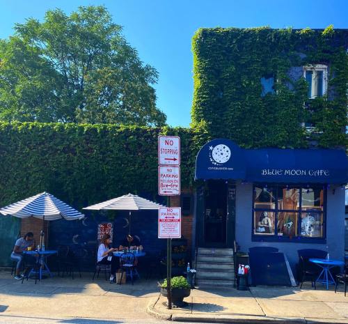 A cozy café with outdoor seating, surrounded by greenery and blue skies, featuring a sign that reads "Blue Moon Café."