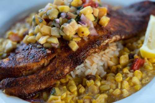 Grilled fish fillet topped with fruit salsa, served over rice and corn, garnished with a lemon wedge.