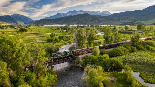 A train crosses a bridge over a river, surrounded by lush greenery and mountains under a cloudy sky.