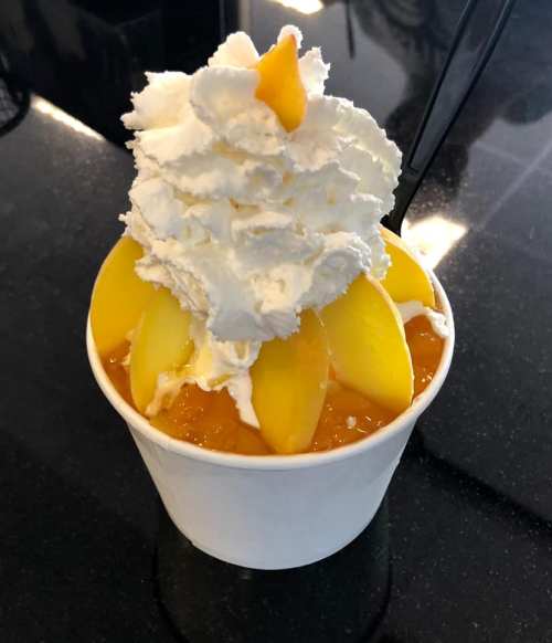 A cup of dessert topped with whipped cream and slices of yellow peaches on a glossy black surface.