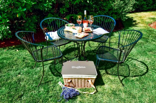 A cozy outdoor picnic setup with a table, chairs, wine glasses, food, and a picnic basket on a grassy lawn.