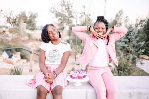 Two smiling girls sit on a ledge, one in a pink outfit and the other in a graphic tee and skirt, with cupcakes nearby.