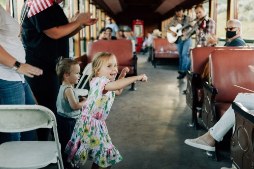 A young girl dances joyfully in a train car while people clap and enjoy live music.