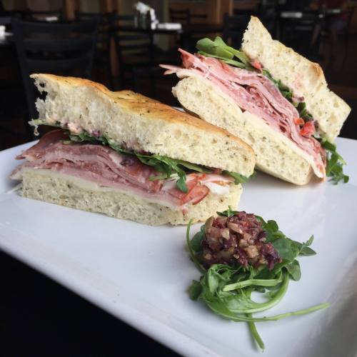 A sandwich with layers of ham, cheese, and greens, served on a plate with a small side of chopped olives.