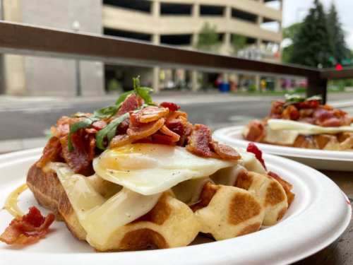 A close-up of a waffle topped with melted cheese, a fried egg, and crispy bacon, served on a paper plate.