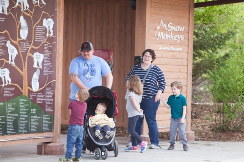 A family with children and a stroller stands outside a wooden structure with a sign about snow monkeys.