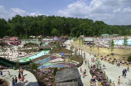 A vibrant water park scene with pools, slides, and crowds enjoying a sunny day surrounded by greenery.