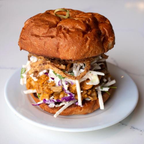 A hearty sandwich with pulled chicken, coleslaw, and crispy onions on a toasted bun, served on a white plate.