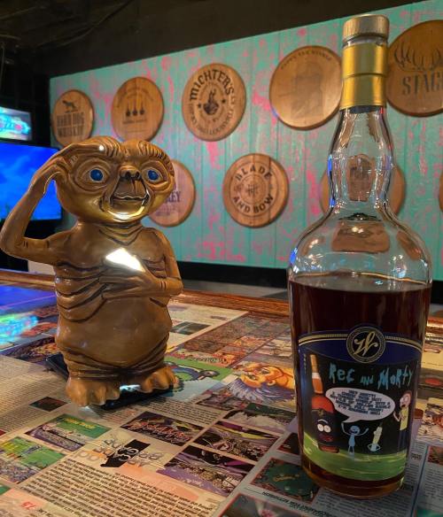 A quirky figurine salutes next to a bottle of "Rick and Morty" whiskey, set against a colorful bar backdrop.