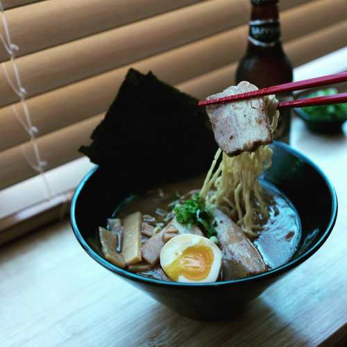A bowl of ramen with noodles, pork, egg, and seaweed, held by chopsticks, with a beer bottle in the background.