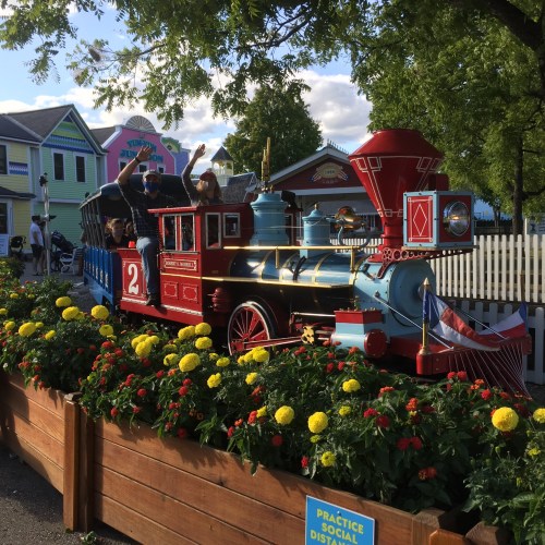 A colorful miniature train with passengers waving, surrounded by vibrant flowers and a cheerful amusement park backdrop.