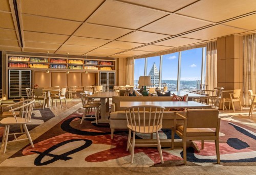 A modern, stylish dining area with large windows, wooden furniture, and a colorful rug, offering a city view.