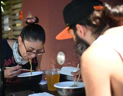 A woman and a man eat noodles from bowls at an outdoor table, with a drink beside them.