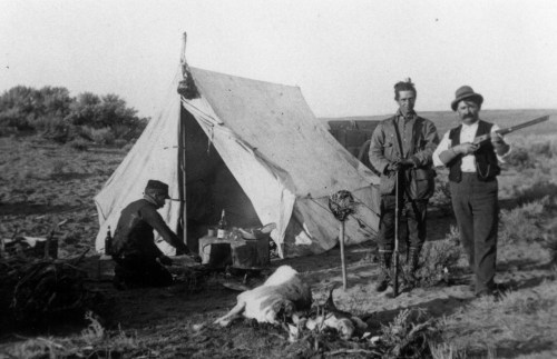 Two men stand by a tent in a desert landscape, one holding a rifle, while another sits nearby with a dog.