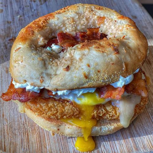 A toasted bagel sandwich with crispy bacon, fried egg, and creamy sauce, oozing cheese on a wooden cutting board.
