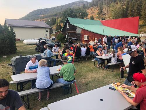 A lively outdoor gathering with people dining at picnic tables near a rustic building and mountains in the background.